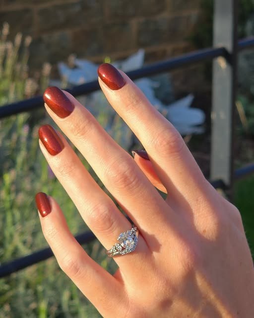 Glossy burnt-orange almond gel nails with a diamond ring, in garden light